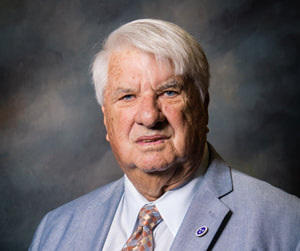 Commissioner Dale Overton appears solemn in front of a shaded dark background wearing a light gray suit coat with lapel pin, white collared button down shirt, and a multicolored spotted necktie.