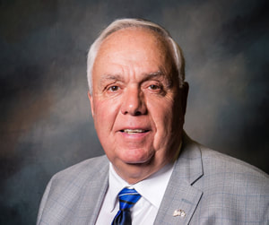 Commissioner David Hawks smiles in front of a shaded dark background wearing a light gray plaid suit coat with lapel pin, white collared button down shirt, and a bright blue necktie with a yellow stripe on the knot.