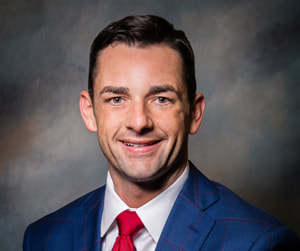 Mayor Bynum smiles before a dark shaded multicolored background wearing a blue suit, white collared button down shirt, and a bright red tie.
