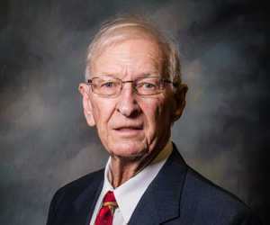 Commissioner Bobby Dunlap is solemn in front of a shaded dark background wearing a black suit coat, white button down collared shirt, and a red and gold necktie.