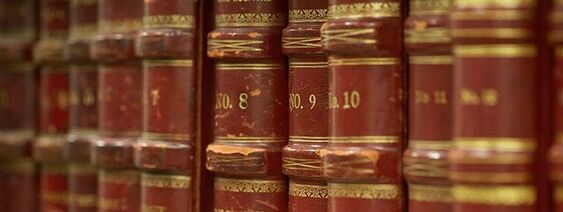 A closeup picture of the middle third of the spines of about eleven volumes of old archived books, titled No. 8, No. 9, No. 10, with other volumes' titles' blurred.  The books are a dark red with shiny gold writing as well as gold detail and filigree embossing.
