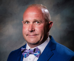 Commissioner Gary Eddings smiles in front of a shaded dark background wearing a bright indigo suit coat, a white collared button down shirt, and a purple and blue striped bowtie.