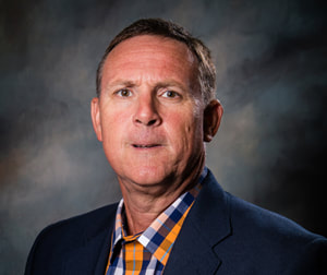 Commissioner Jack Vincent appears solemn in front of a shaded dark background wearing a dark navy suit jacket and a orange, navy, and white plaid button down collared shirt. 