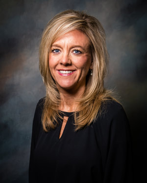 With medium length blonde hair and blue eyes, County Clerk Kim Hughey smiles before a multi-colored background wearing a black top.