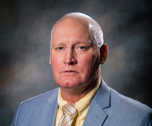 Commissioner Larry Hudson wears a slight smile in front of a shaded dark background wearing a gray suit coat, a pastel yellow collared button down shirt, and a white and shades of gold shiny and striped print necktie.