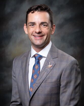 Mayor Bynum smiles in front of a gray backdrop, eyes shining with coal black hair, a snappy blue suit, a solid red tie and a white button down collared shirt.