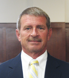 Sheriff Mike Wilson stand before a wood paneled background in a navy suit, crisp white shirt, and yellow and white striped tie.